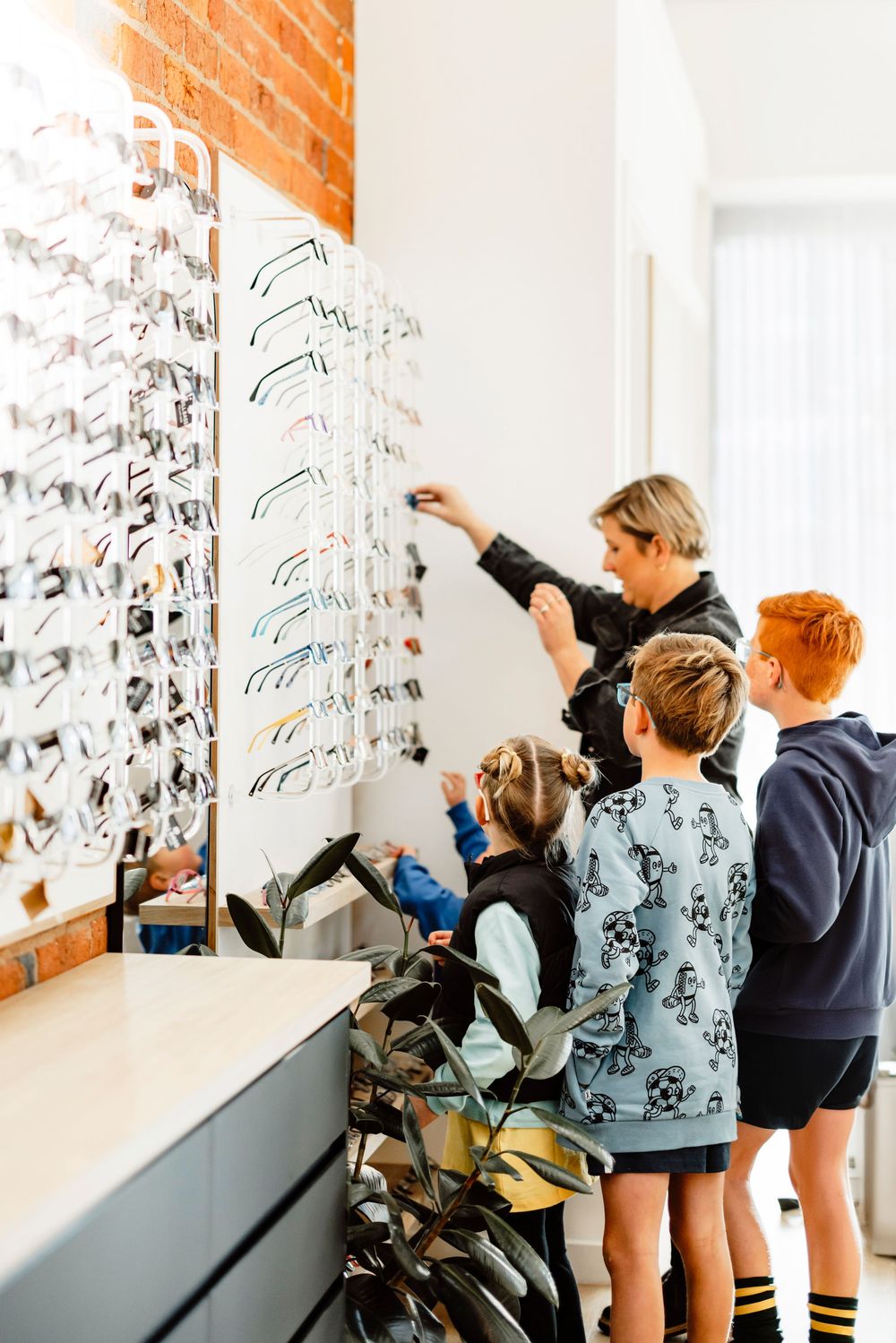 Woman helping three children select eyeglasses from a display wall in an optical store.