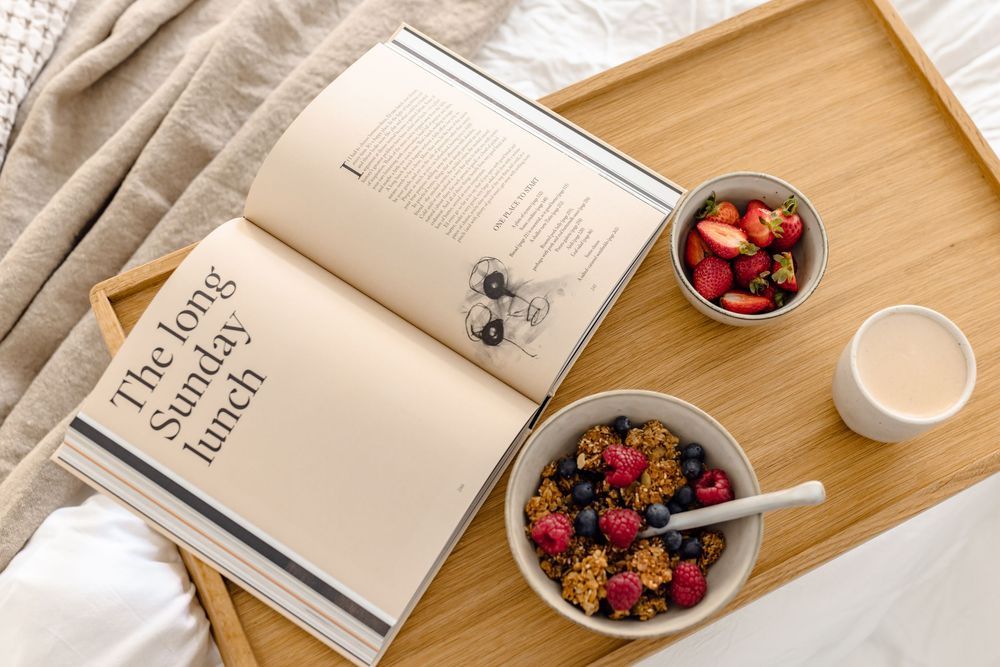 Breakfast tray in bed with granola, fresh berries, coffee, and an open cookbook.