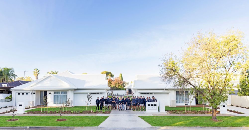 Large white ranch-style home with group of people posing on front lawn surrounded by lush green landscaping.