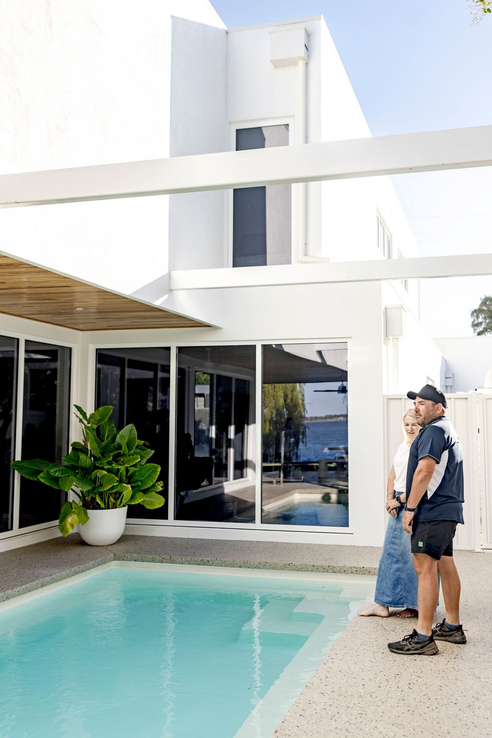 Couple standing beside a modern white two-story home with a sleek rectangular pool and tropical plant.