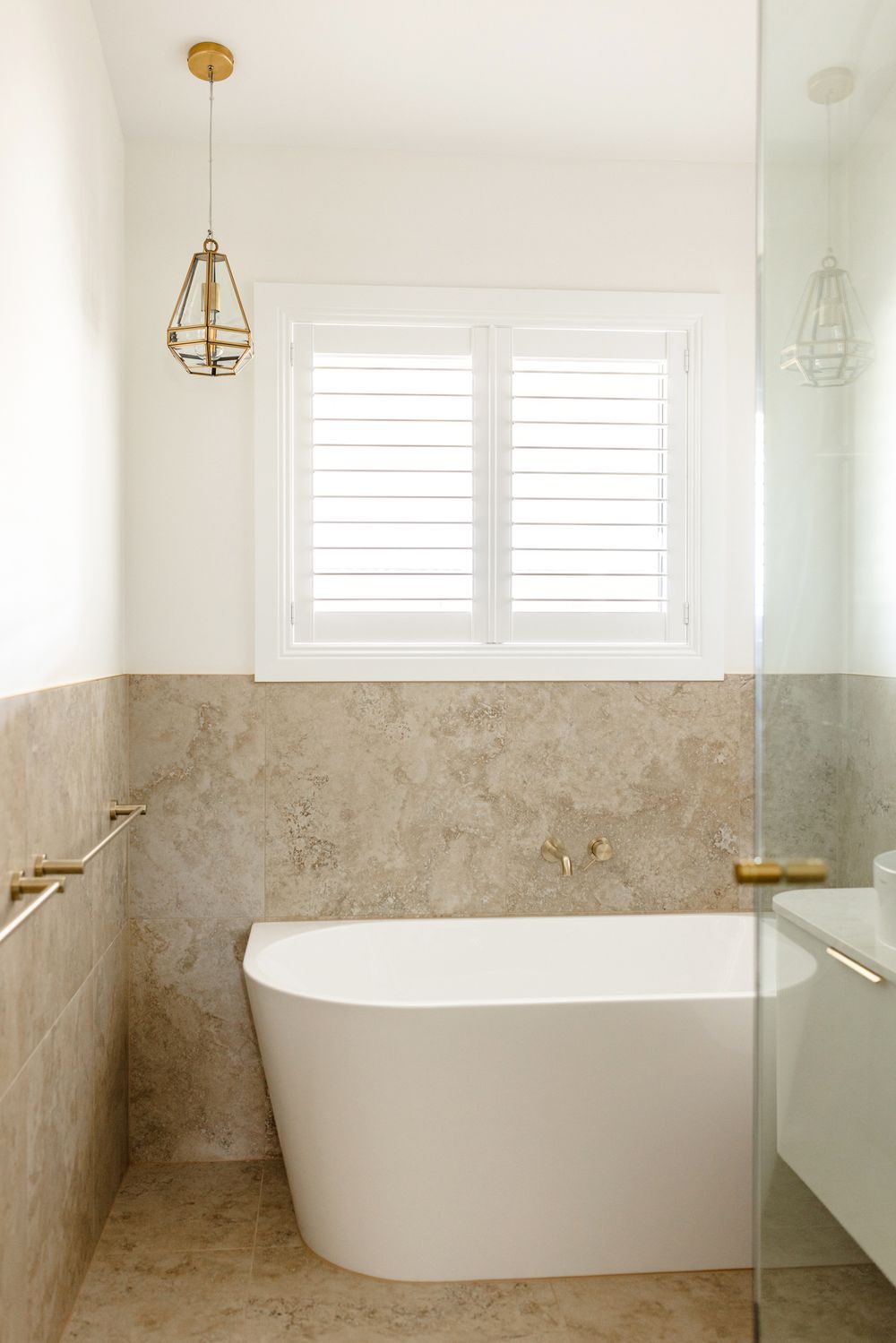Luxurious bathroom with freestanding white bath, travertine tiles, gold fixtures and pendant light.