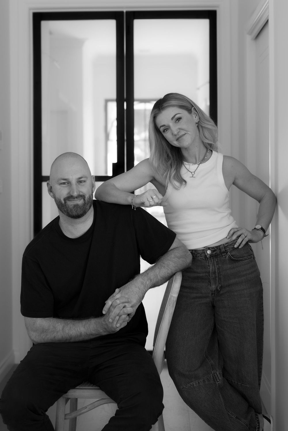 Black and white portrait of bald bearded man and blonde woman posing in bright hallway.