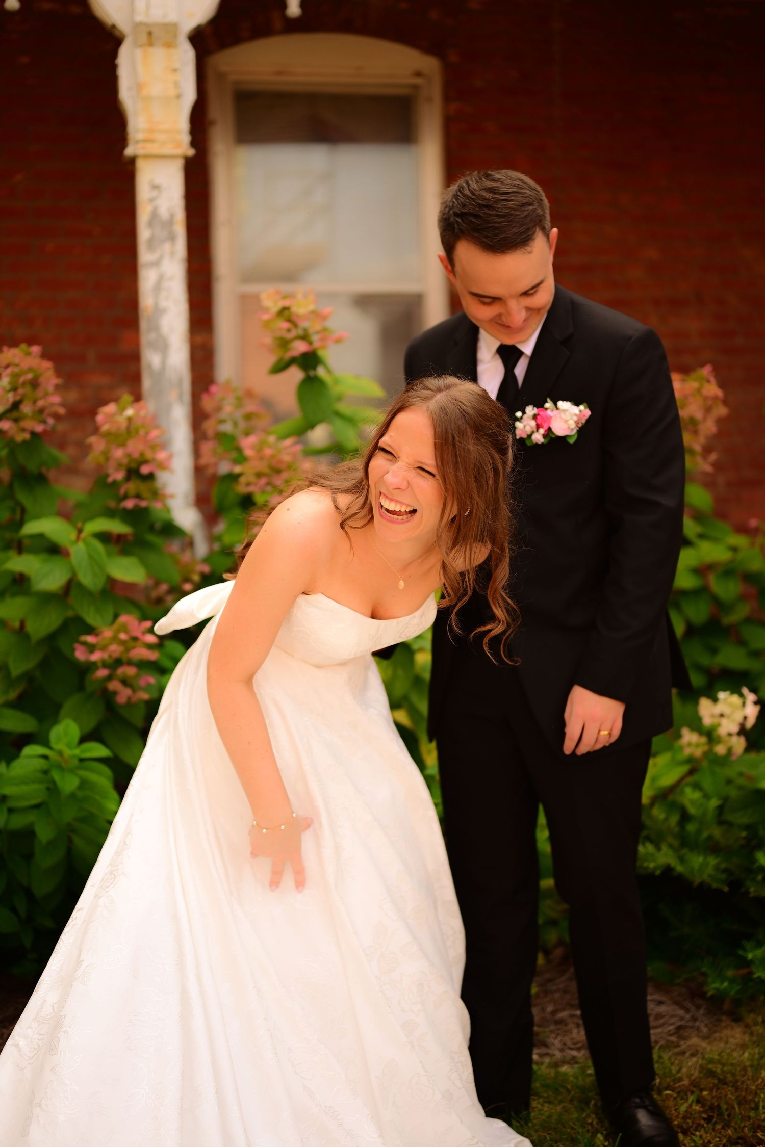 Newlywed couple shares joyful candid moments in a garden setting with blooming flowers and columns.