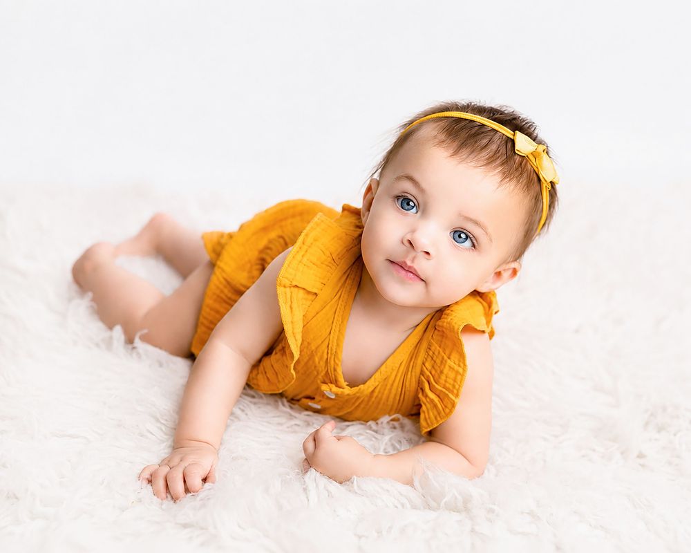 Adorable baby in yellow romper lying on white fluffy blanket, looking up with bright blue eyes.