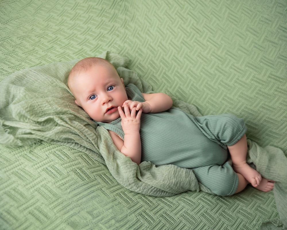 Newborn baby in green outfit lying on green textured blanket with pillow, peaceful infant portrait photography.