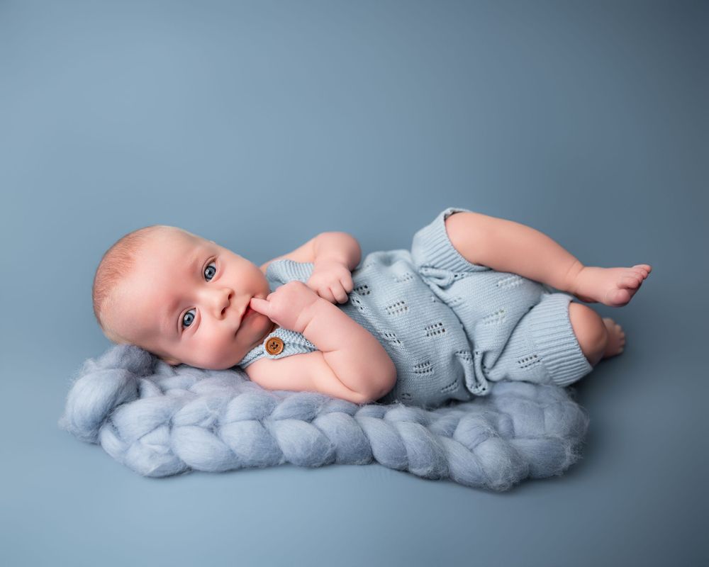 A newborn baby in a blue knit romper lies on a soft blue merino wool braid against a blue backdrop.