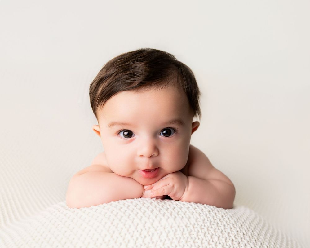 Adorable baby with dark hair resting chin on hands, lying on a white knit blanket against a soft white background.
