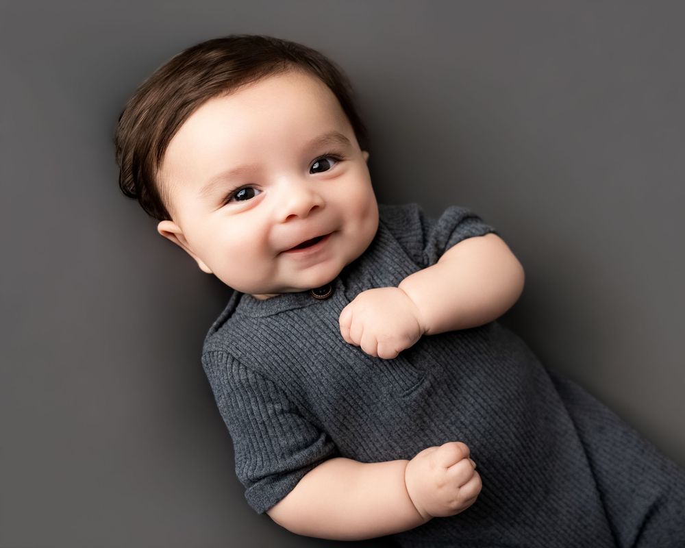 Smiling baby boy in gray outfit lying down, professional newborn portrait photography on gray background.