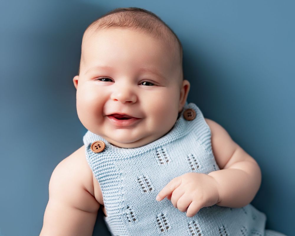 Smiling baby in a light blue knit romper lying on a blue background, looking happy and cheerful.
