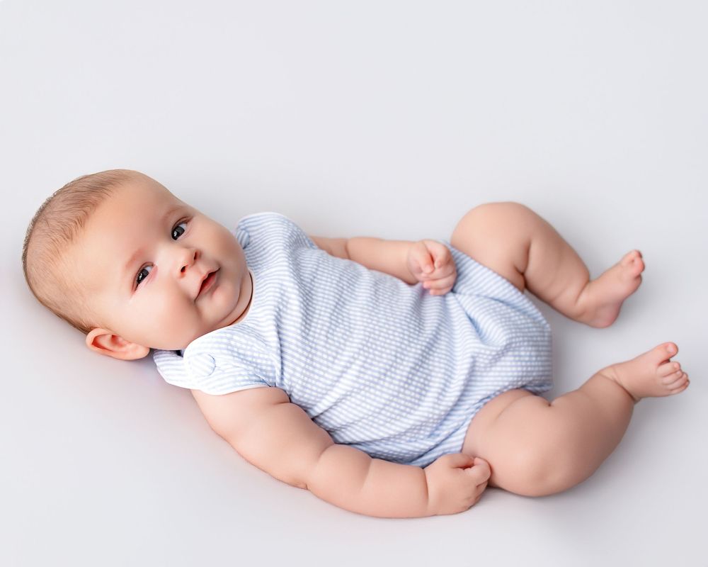 Smiling baby in light blue striped onesie lying on white background, looking up with bright eyes.