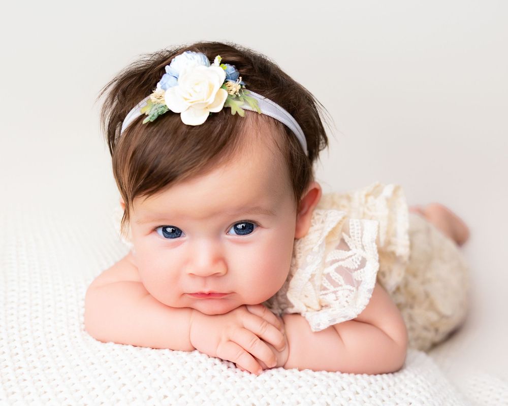 Adorable baby girl with blue eyes wearing a floral headband and lace romper, lying on a white blanket.