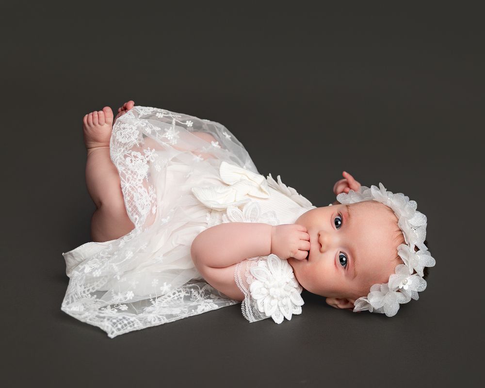 Adorable baby in white lace dress and floral headband lying on gray background.