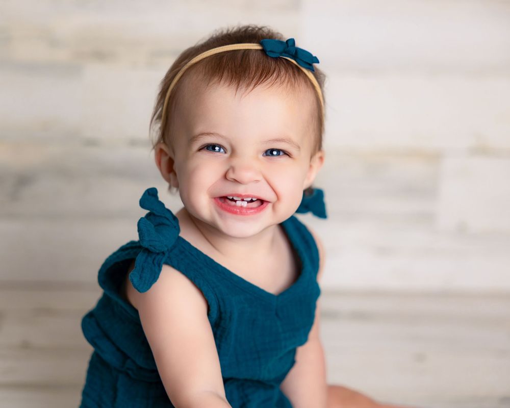 Smiling baby girl in teal ruffle romper with bow headband poses for a professional portrait photo.