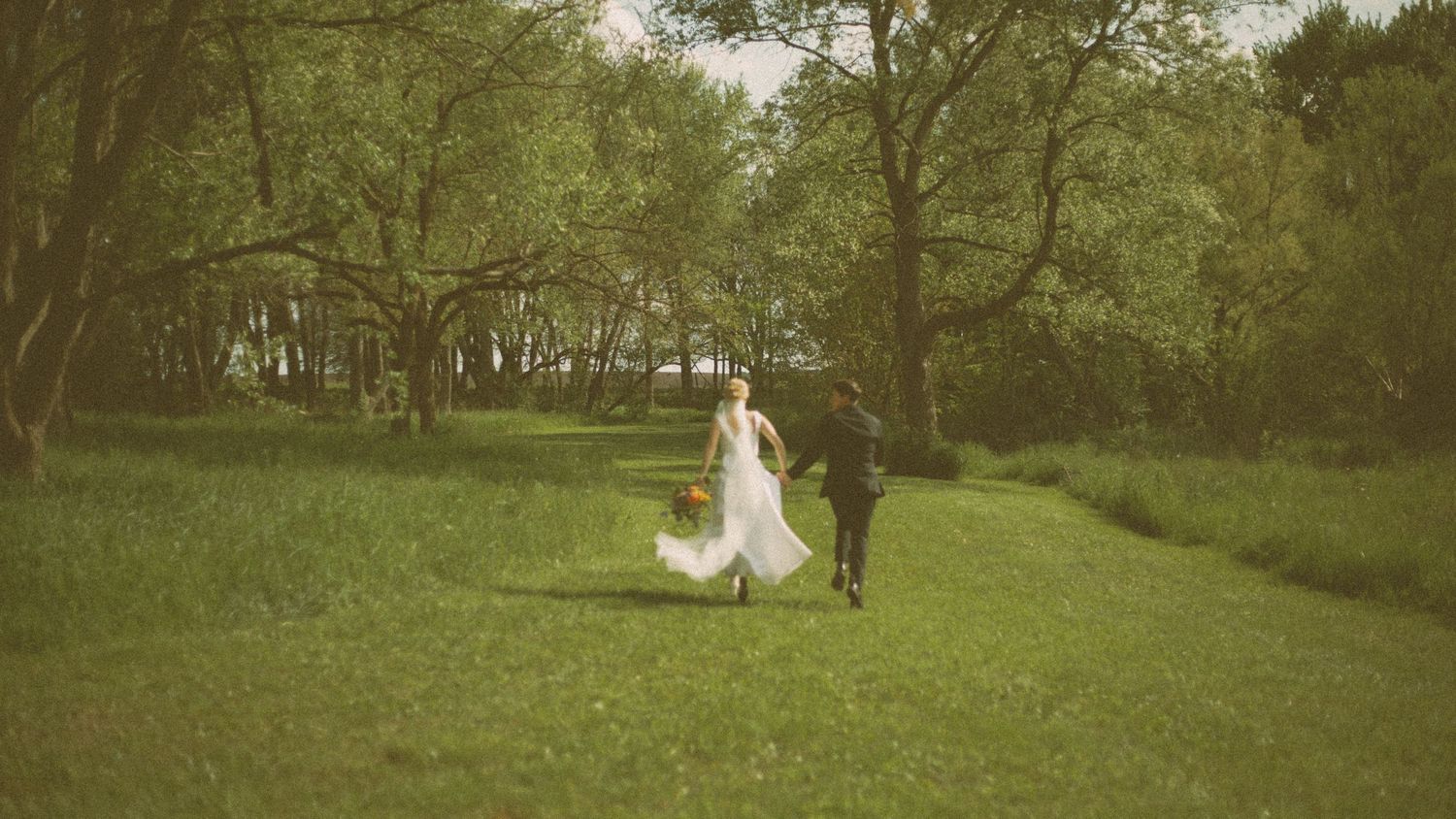 A couple walks through a lush green landscape together in vintage-style wedding attire.