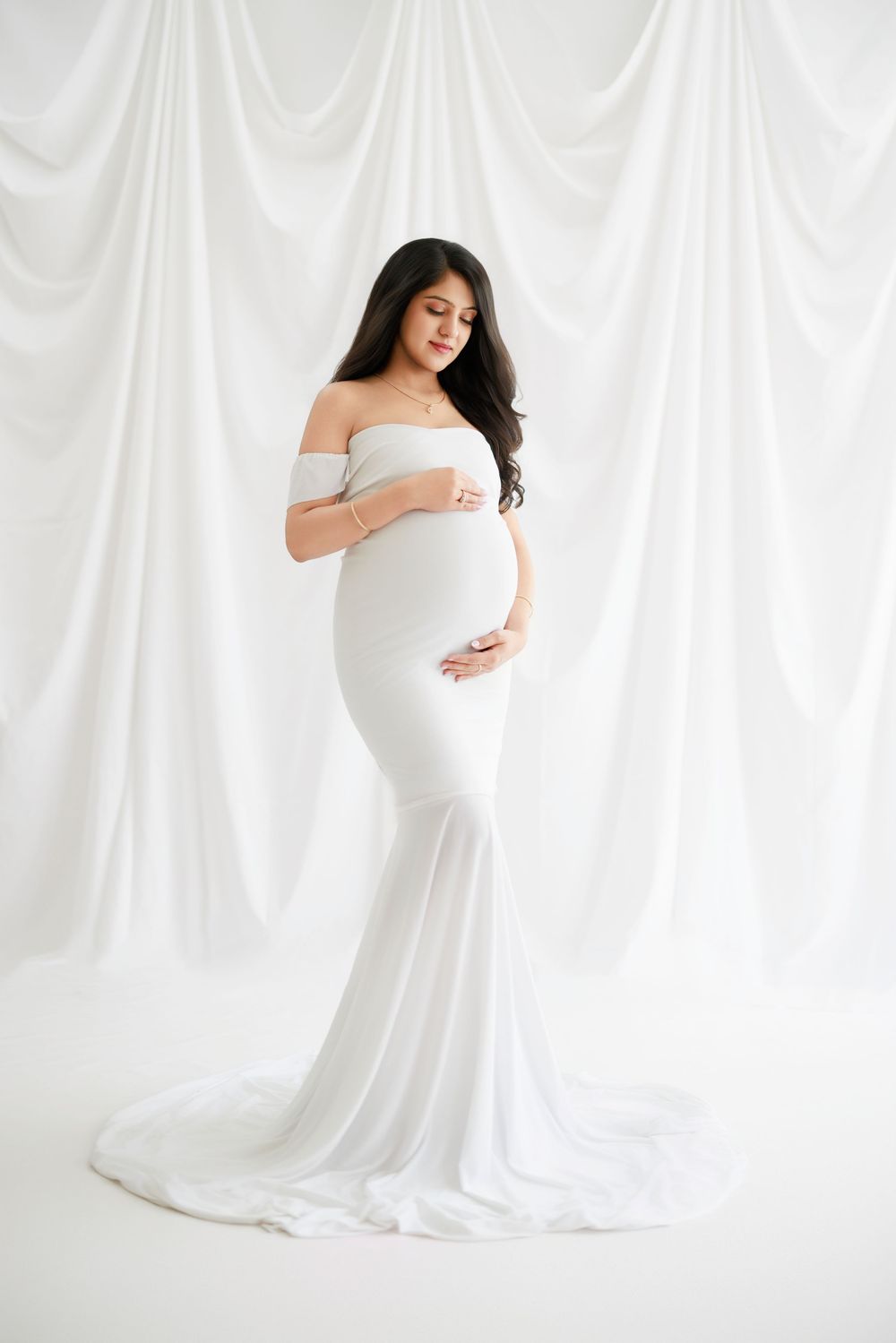 A maternity portrait features an elegant off-shoulder white gown against a soft white draped backdrop in a minimalist style.