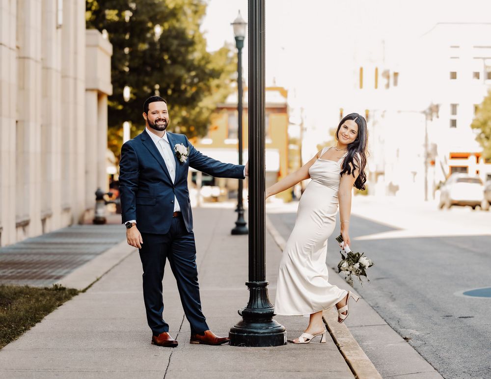 Newlyweds strike playful poses on a city sidewalk next to an ornate lamppost.