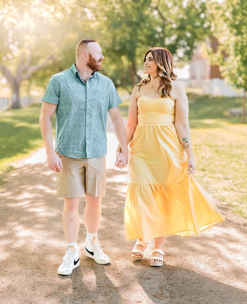 A couple holds hands while walking on a path in a sunny park, wearing yellow dress and green shirt on a warm day.