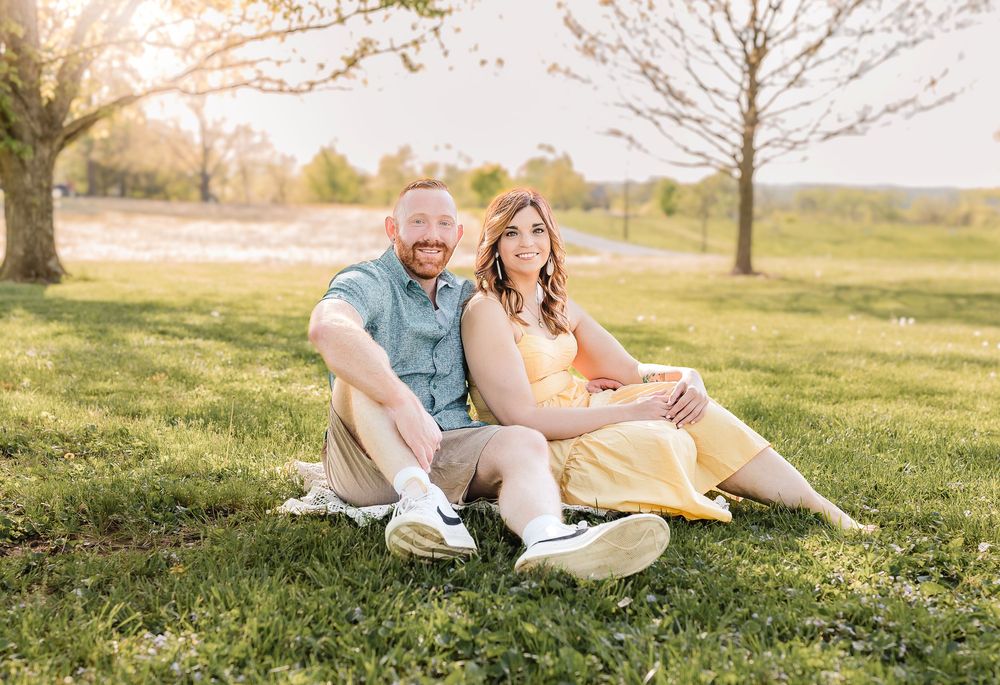 A couple sits together on a blanket in a sunny spring field with trees in the background.