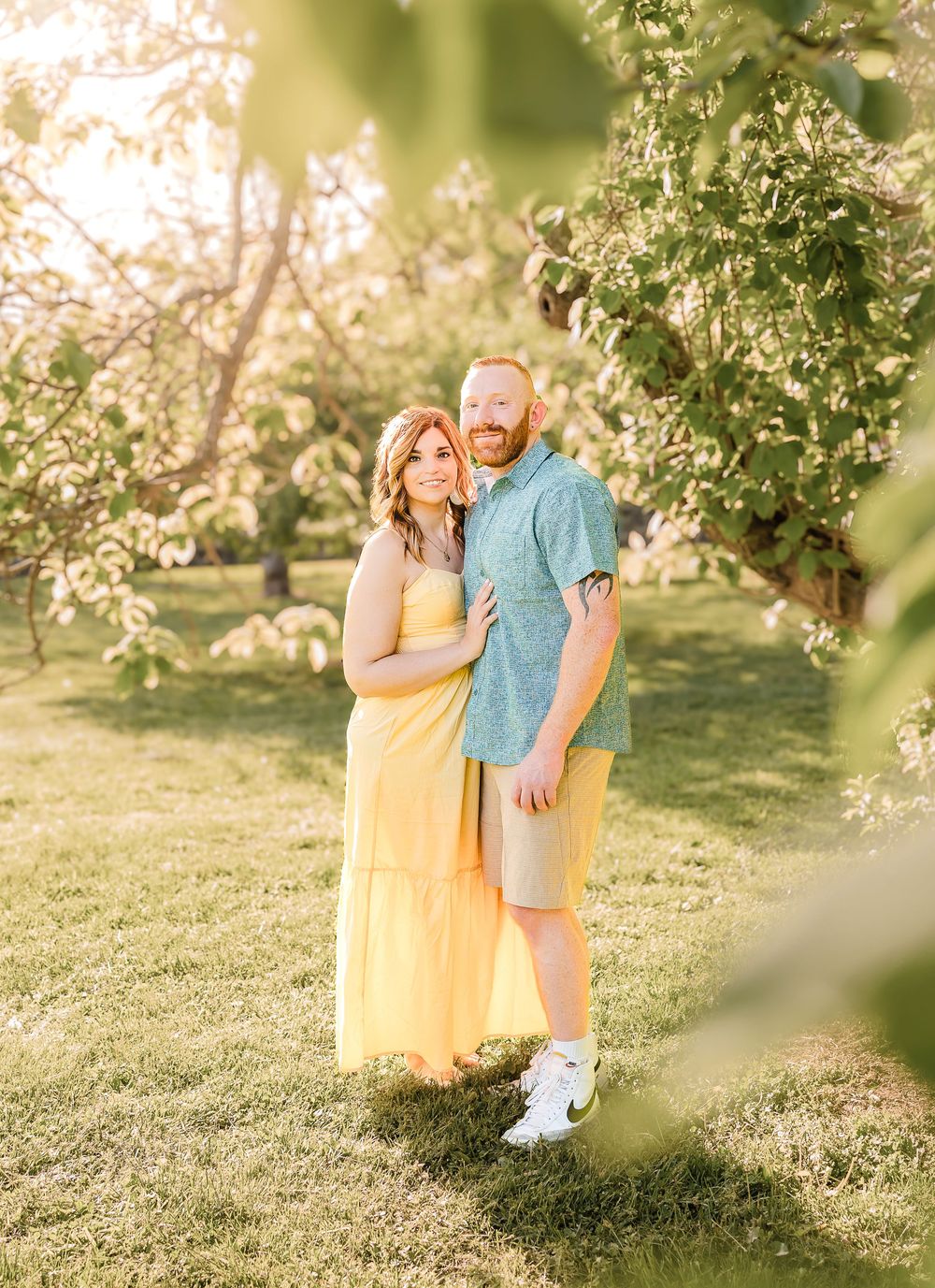 A couple embraces under blooming spring trees in a sunlit garden setting wearing yellow and blue casual attire.