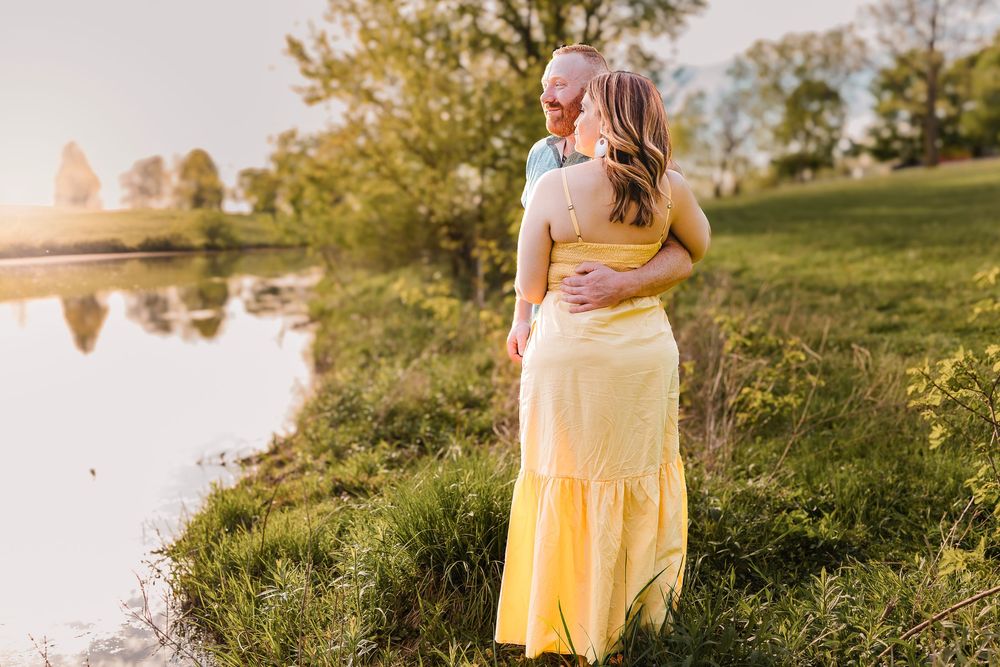 A couple embraces near a tranquil pond at sunset with fall trees reflecting in the water.