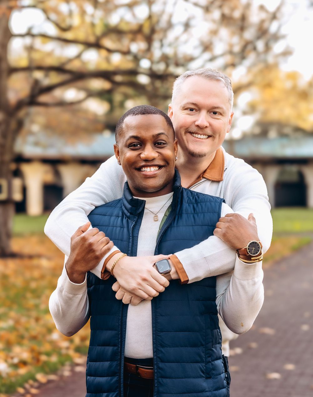 Couple in casual autumn attire sharing a loving embrace outdoors with fall foliage in background.
