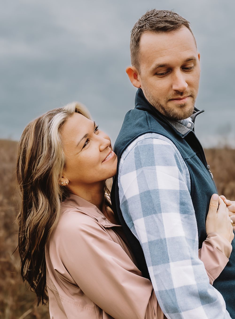 A romantic couple embraces outdoors wearing casual clothing including a plaid button-down shirt and pink sweater.