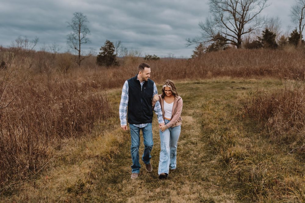 A couple walks hand in hand through a rustic autumn field under moody cloudy skies.