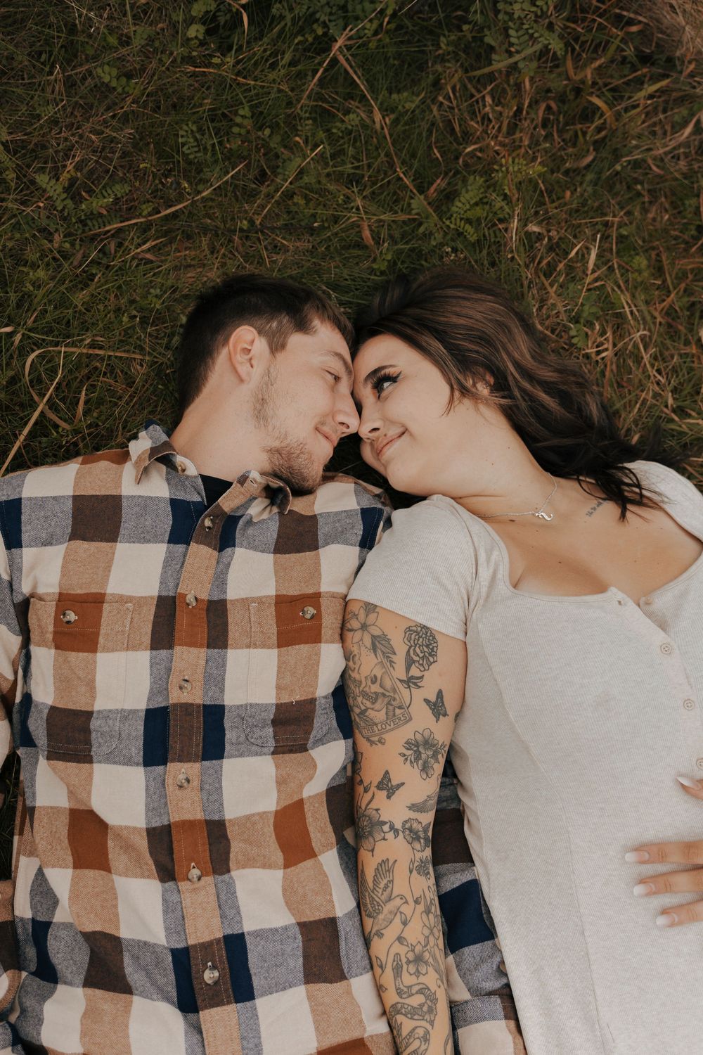 A romantic couple lies together on a plaid blanket in nature, with one person wearing a white shirt and having tattoos.