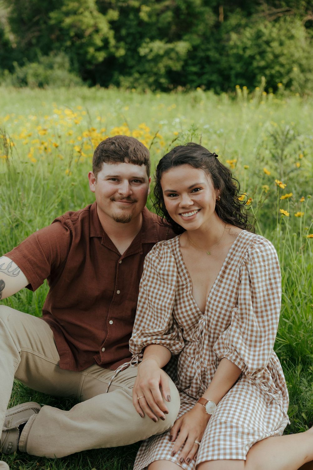 A couple relaxes together in a sunny meadow with yellow wildflowers wearing brown and gingham outfits.