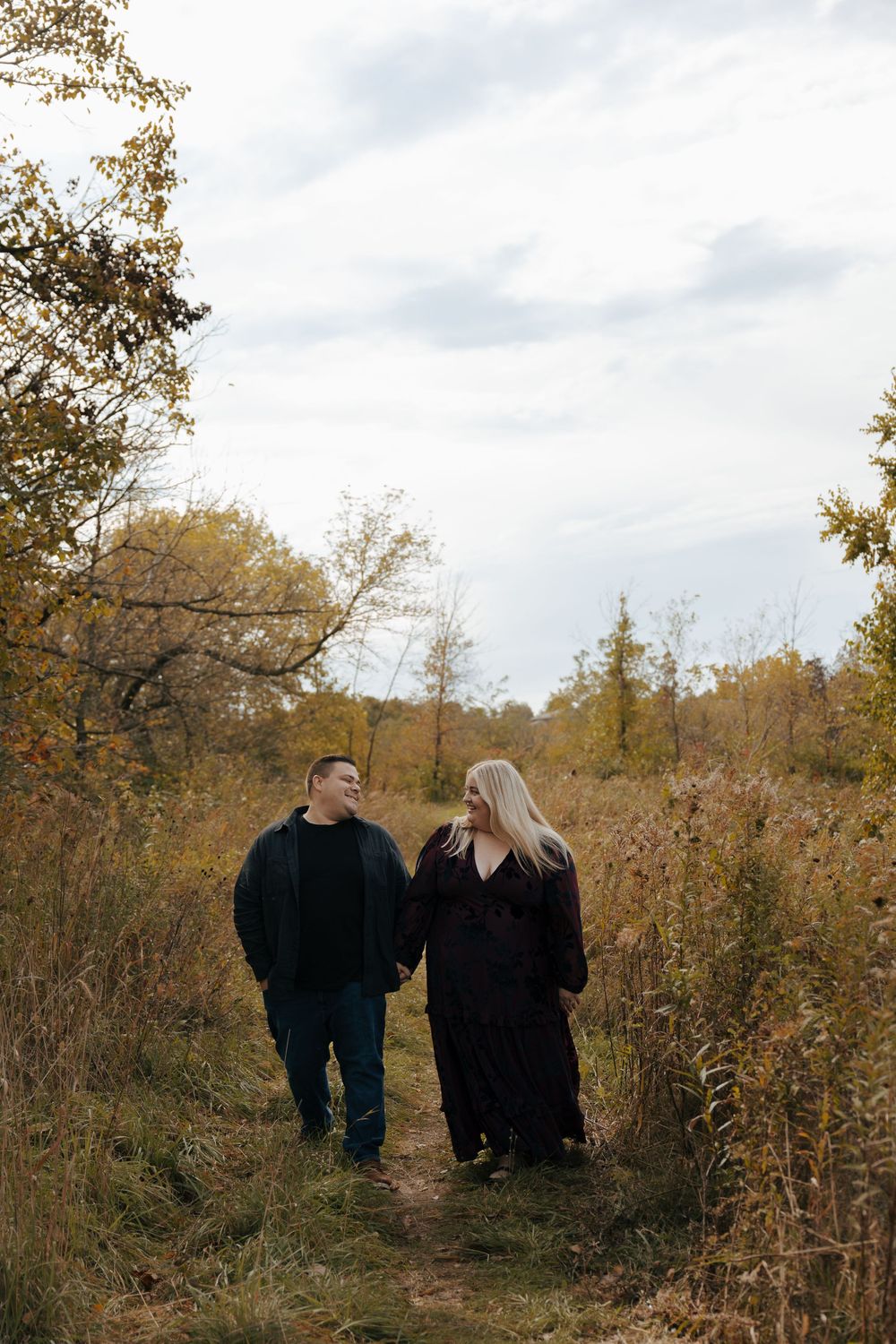 A couple walks together on an autumn trail surrounded by golden foliage and tall grass on a cloudy day.