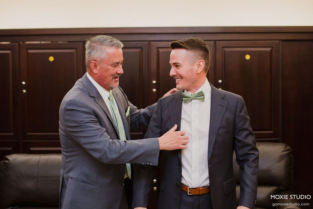 Two people in formal attire share a joyful moment, with matching green bow tie and tie against dark wood paneling.