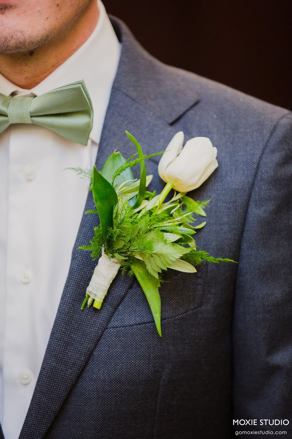 Close-up of white floral boutonniere with greenery pinned to gray suit lapel with mint bow tie.