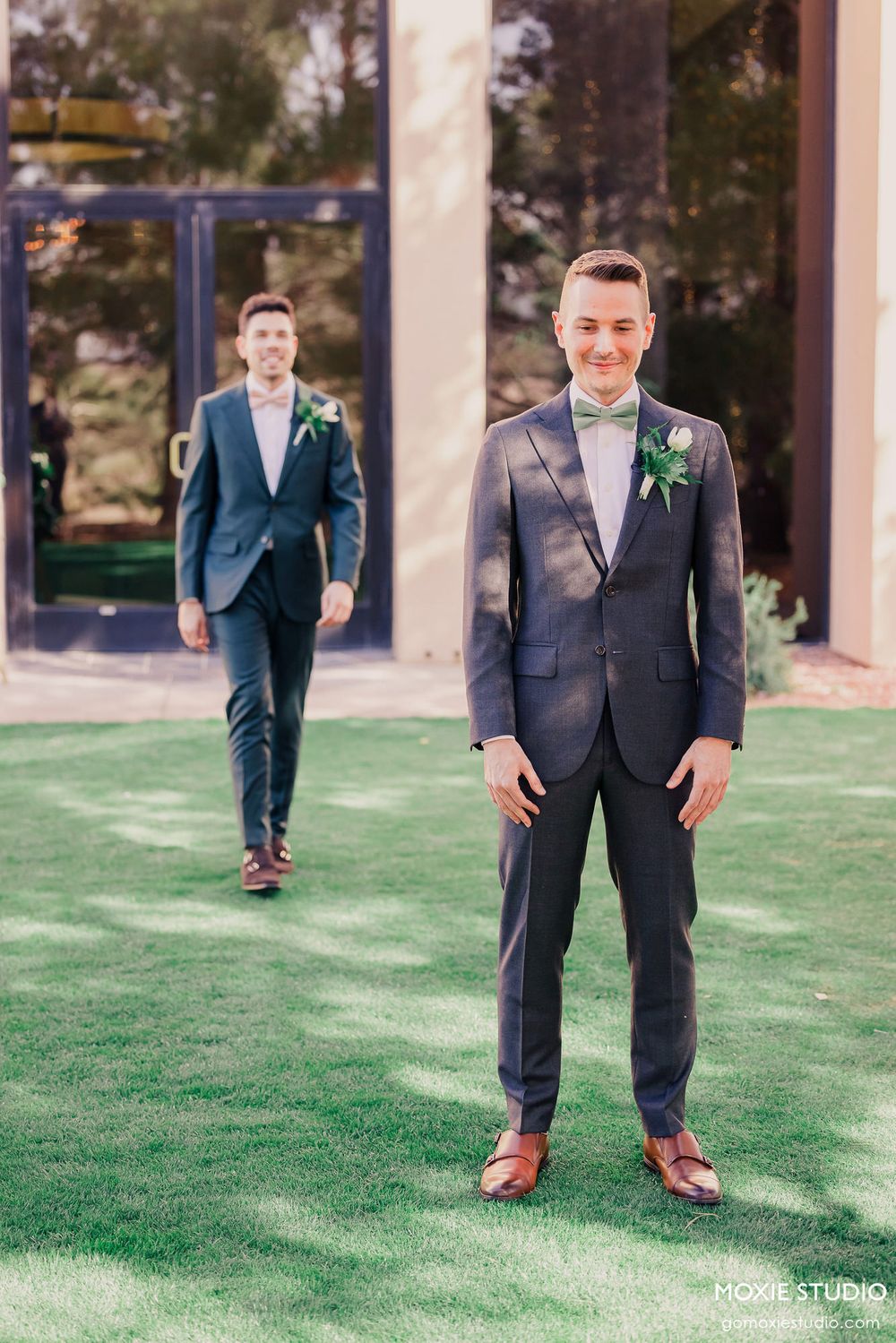 Two groomsmen in navy suits and bow ties stand on manicured lawn during outdoor wedding photoshoot.