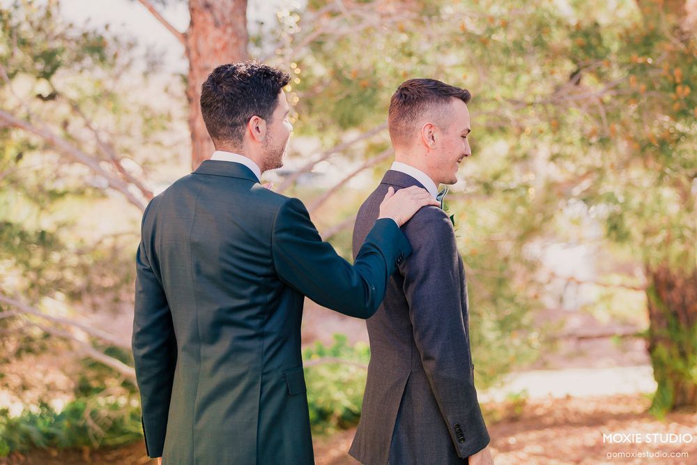 Two people in dark suits share a candid moment outdoors with autumn foliage in the background.