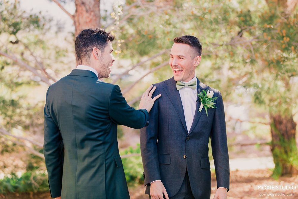 Two people in formal attire laugh together during an outdoor event with trees in the background.