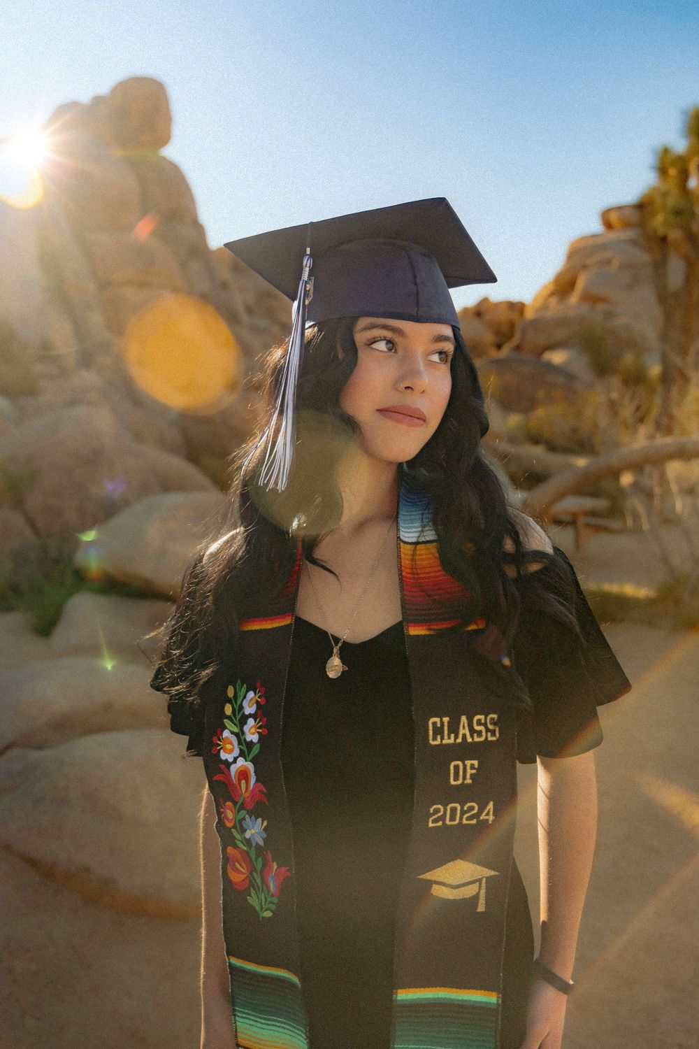 Graduate in cap and gown with Class of 2024 poses at sunset in desert landscape with rocks and lens flare.
