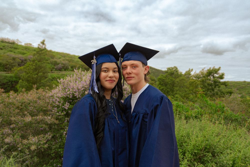 Two graduates in blue caps and gowns stand together outdoors on a hillside under cloudy skies.