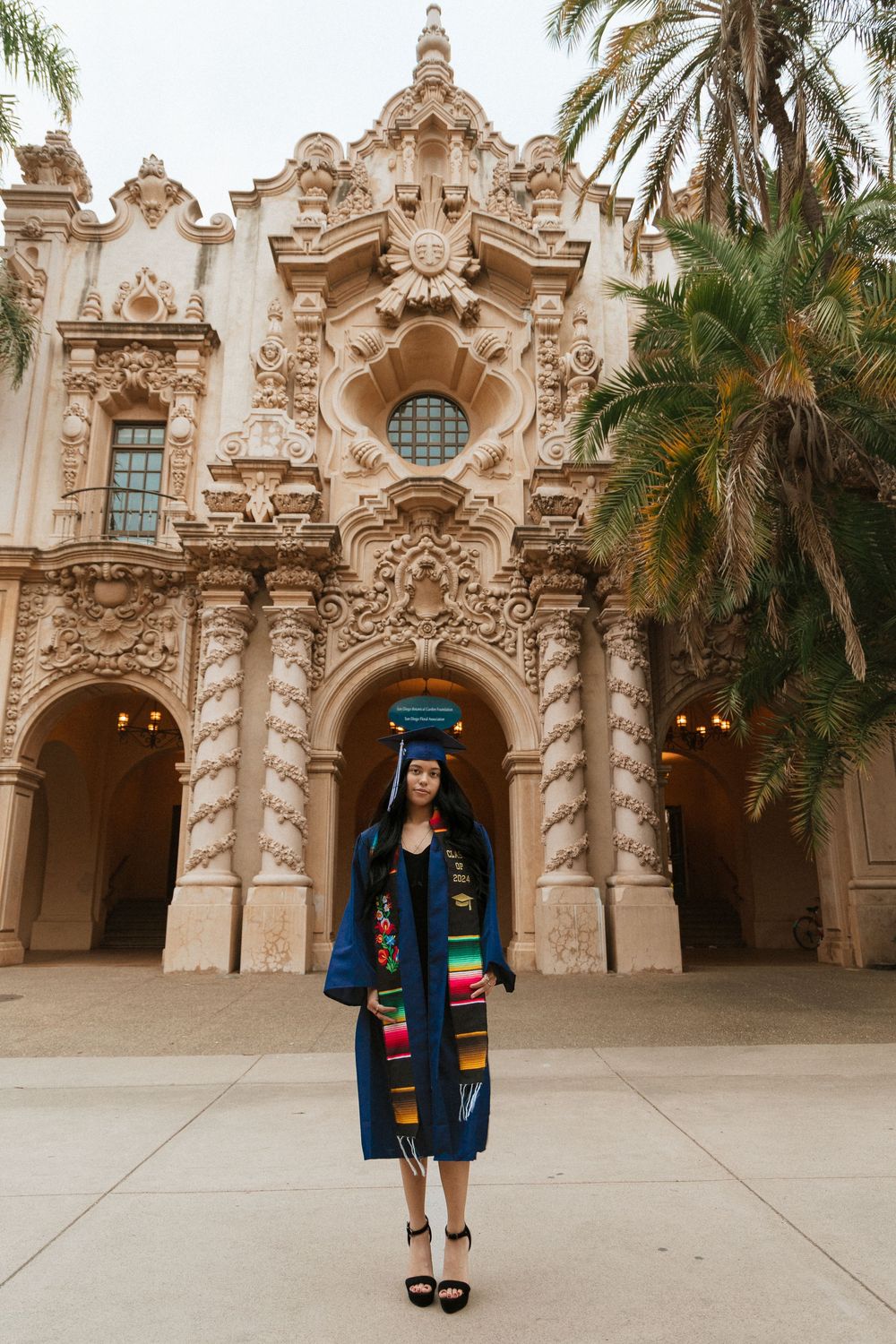 Graduate stands in front of ornate Spanish Colonial architecture at Balboa Park wearing a blue denim jacket and graduation stole.