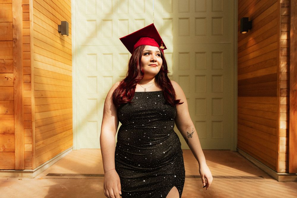 Graduate wearing sparkly black dress stands confidently in a hallway with wooden walls and white doors.