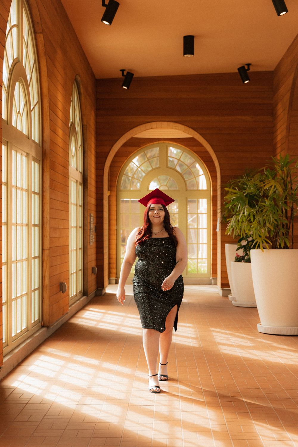 Graduate poses in sunny corridor with arched windows wearing sparkly black dress and red graduation cap.