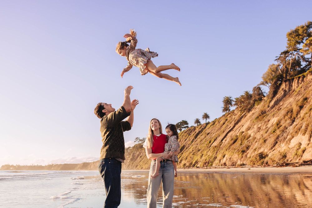 Energetic family playtime with child being tossed in air at beach with cliffs behind.