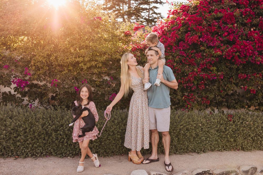 A group enjoys a sunset photoshoot in front of vibrant red bougainvillea flowers along a walkway.