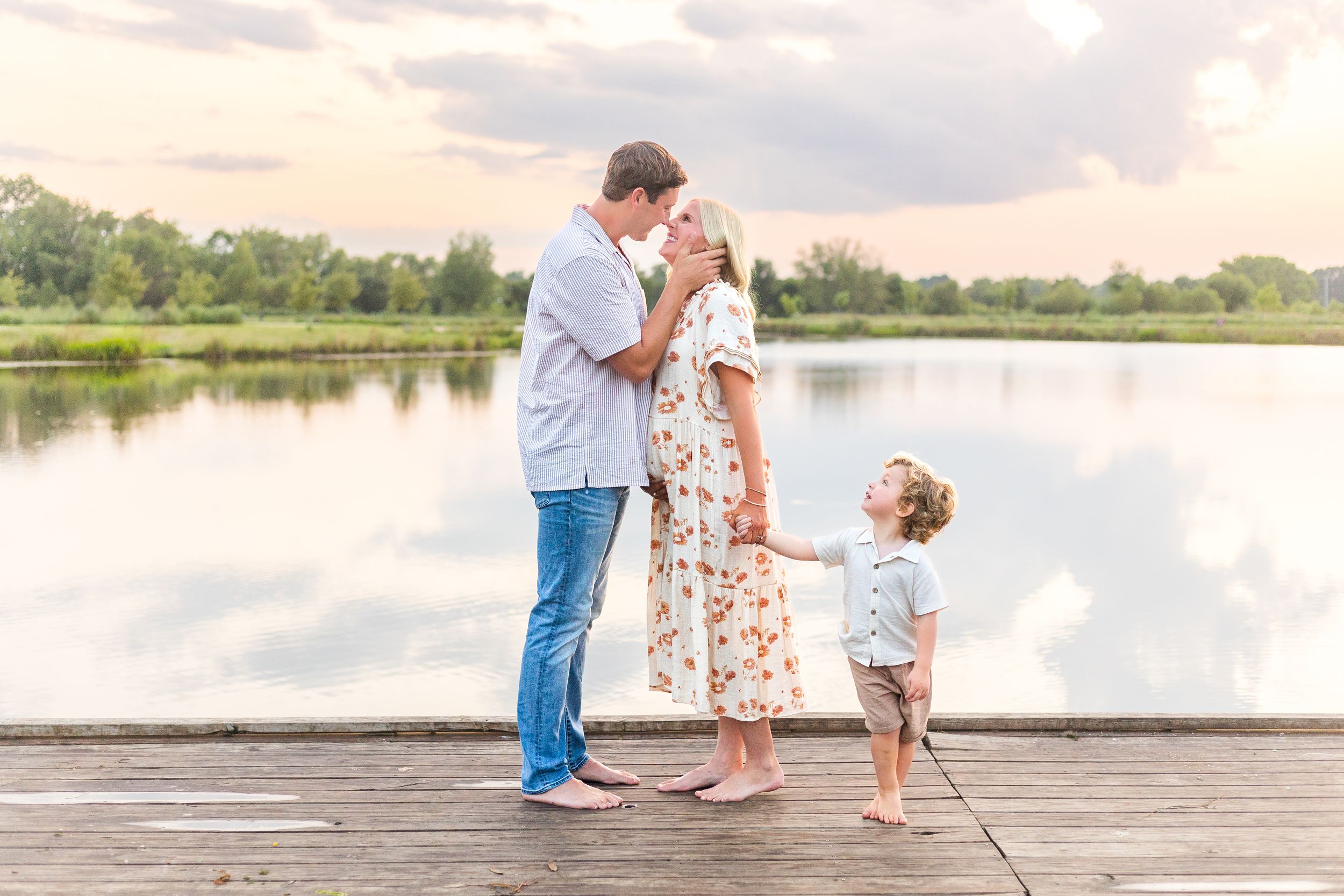 Parents sharing a sweet moment with their young daughter on a wooden dock beside a peaceful lake.