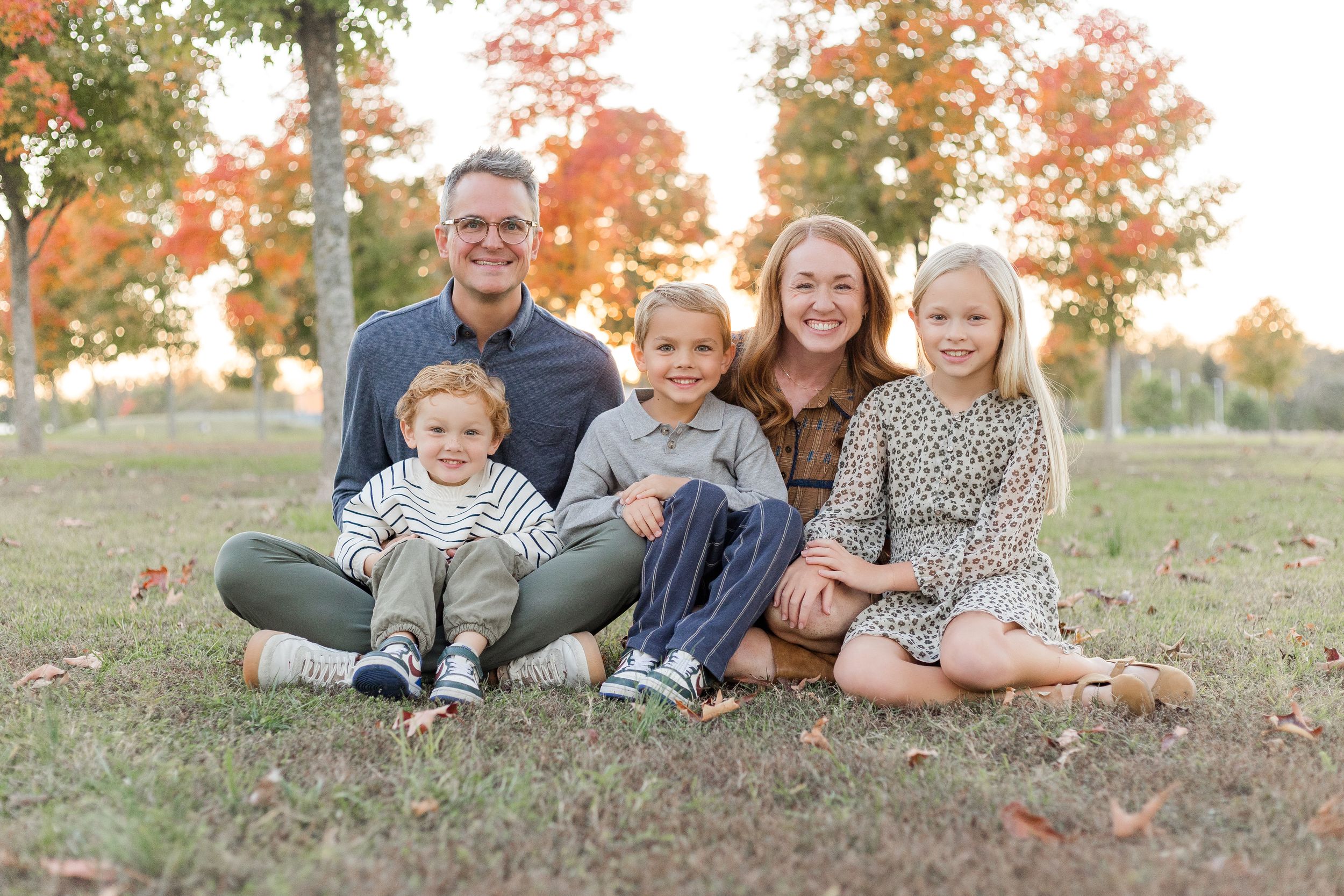 Family of five sitting together on the grass surrounded by colorful autumn trees during an outdoor portrait session.
