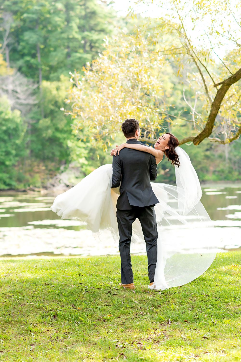 A wedding couple dances playfully on a grassy hill by a river with the bride's veil flowing in the autumn breeze at Springwater Conservation