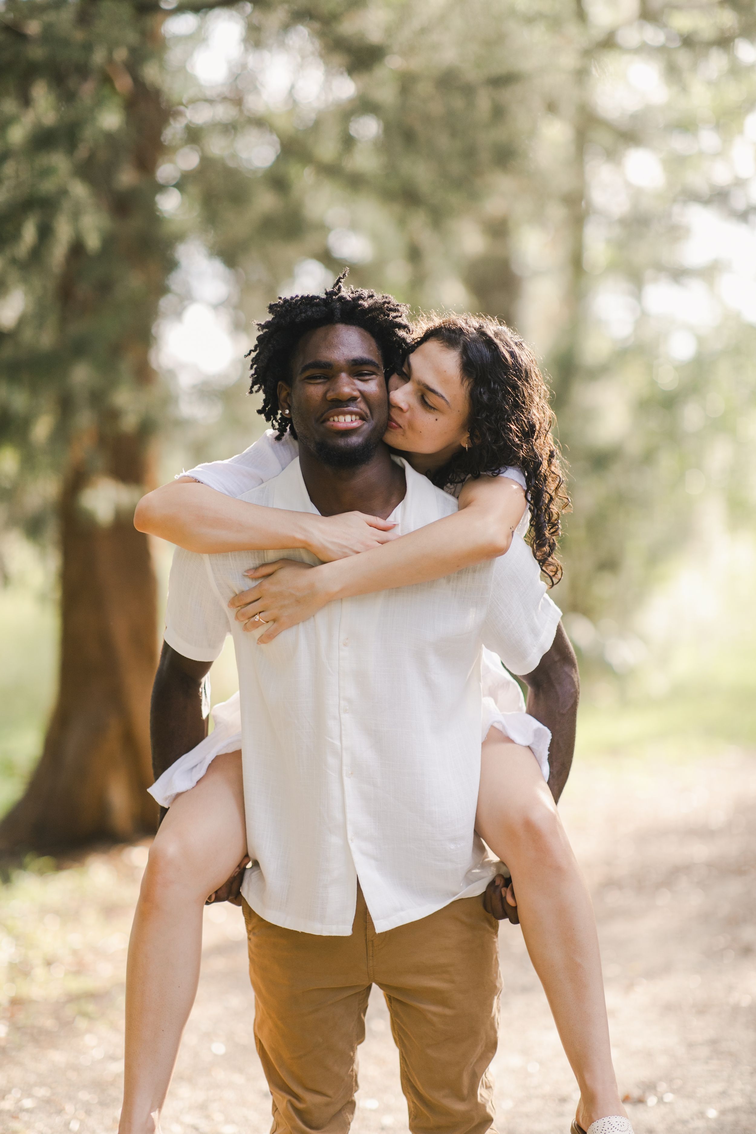 A woman kisses a smiling man giving her a piggyback ride for engagement session, a sunny tree-lined path at park in Beaufort South Carolina.