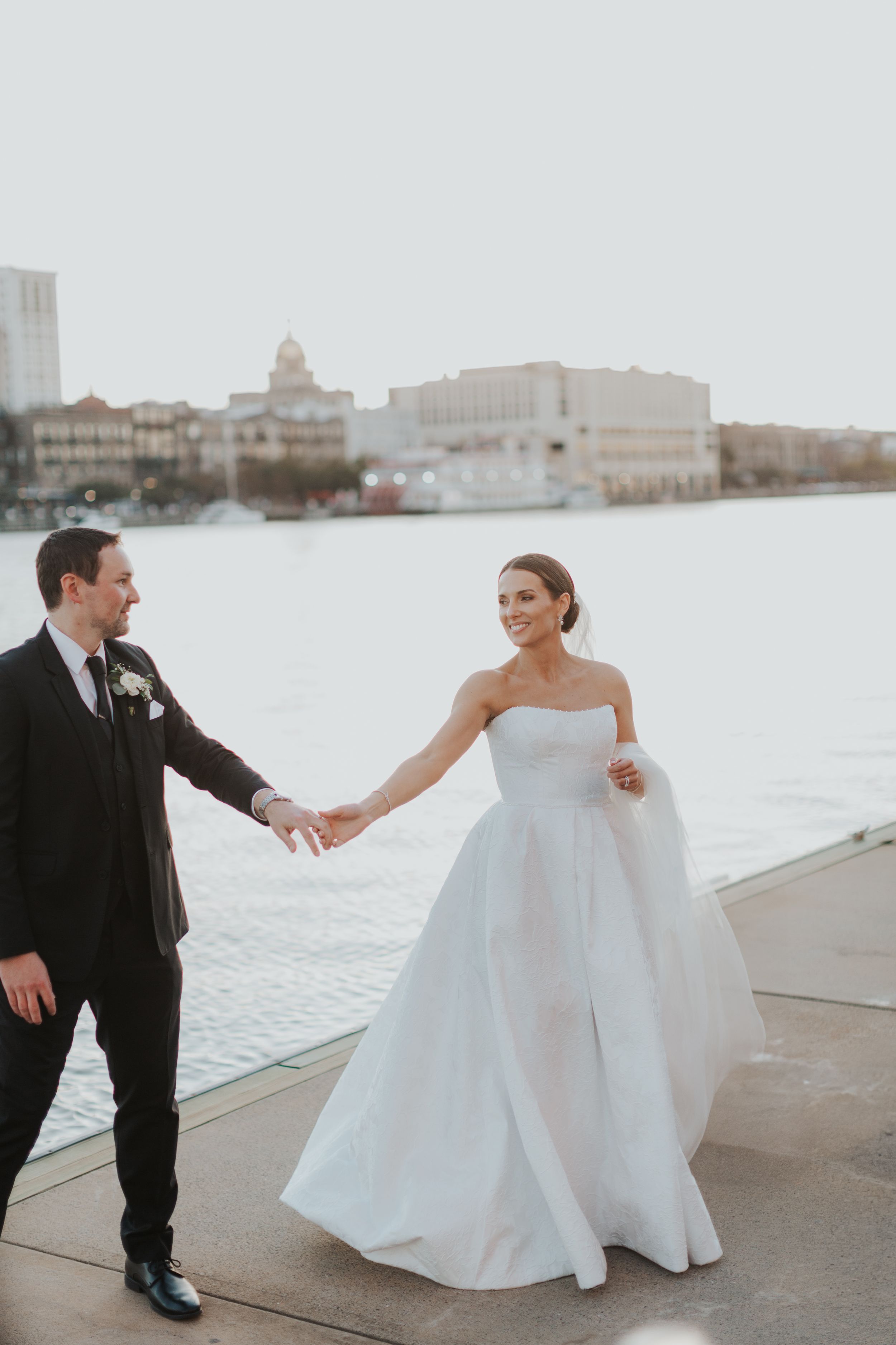 Wedding photography with bride and groom in black suit by waterfront in Savannah, Georgia with city skyline backdrop.