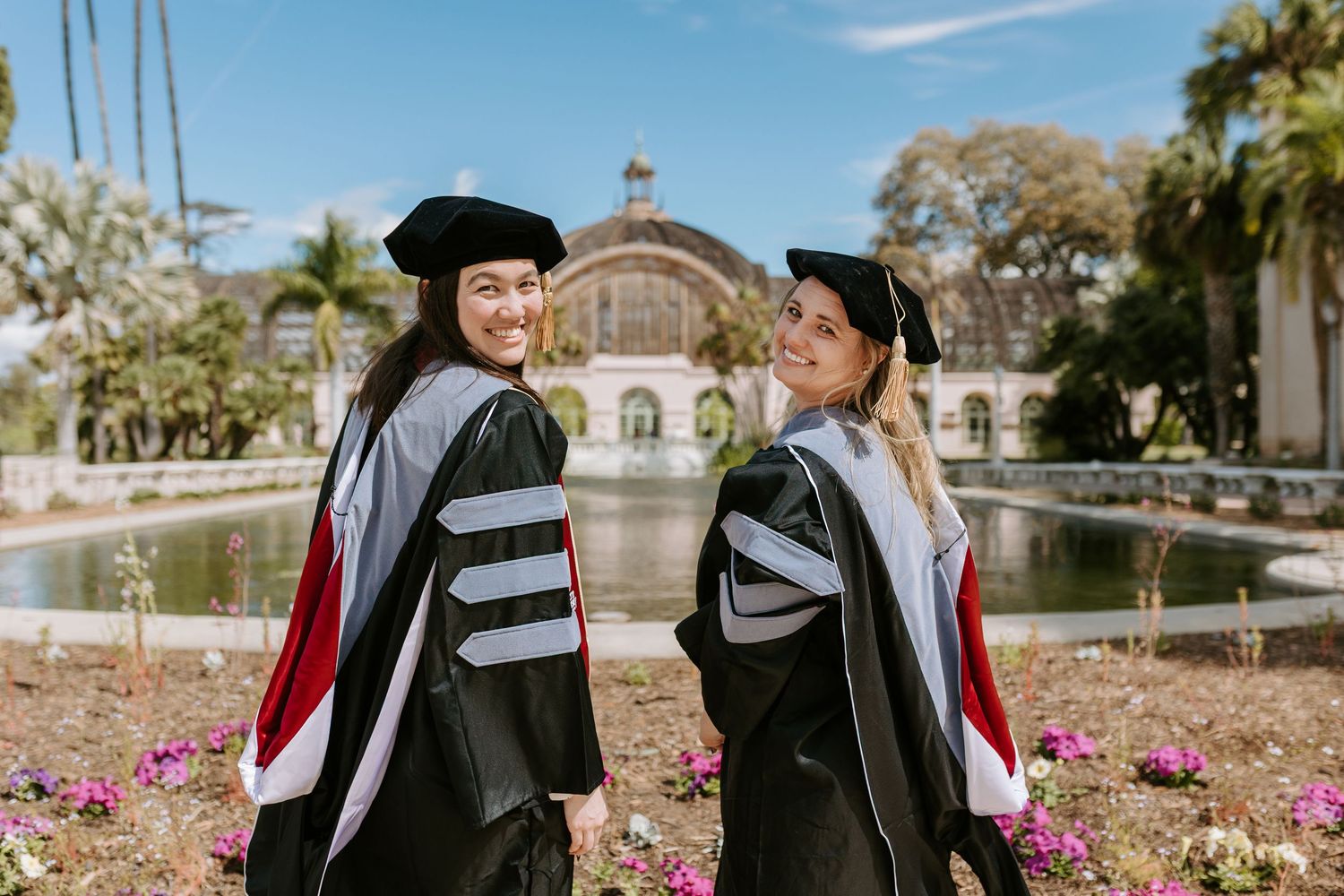 Two doctorate graduates smile in front of the Botanical Building at Balboa Park in San Diego.