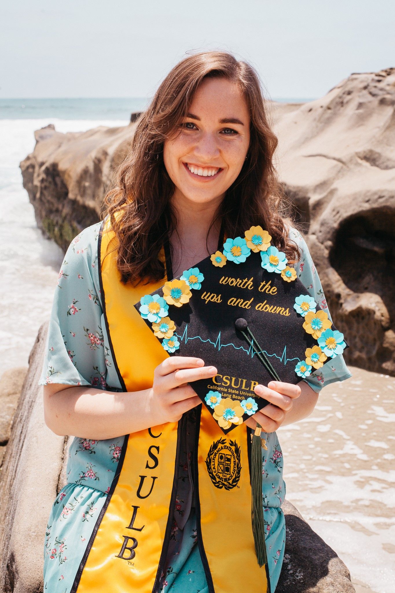 A girl wearing a blue dress and graduation sash holds a decorated graduation cap.