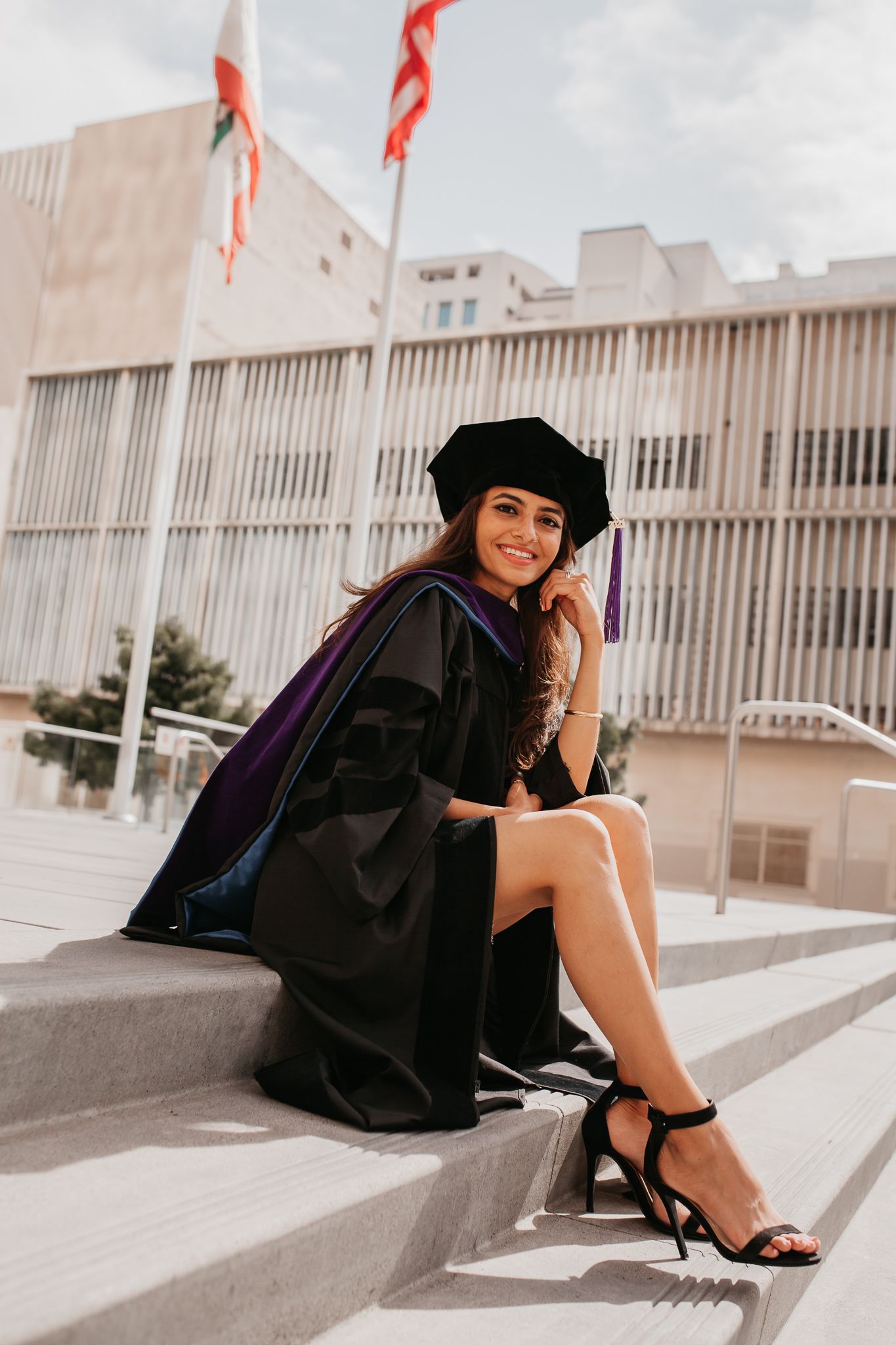 A woman wearing a doctorate cap and gown sits on steps on a sunny day.